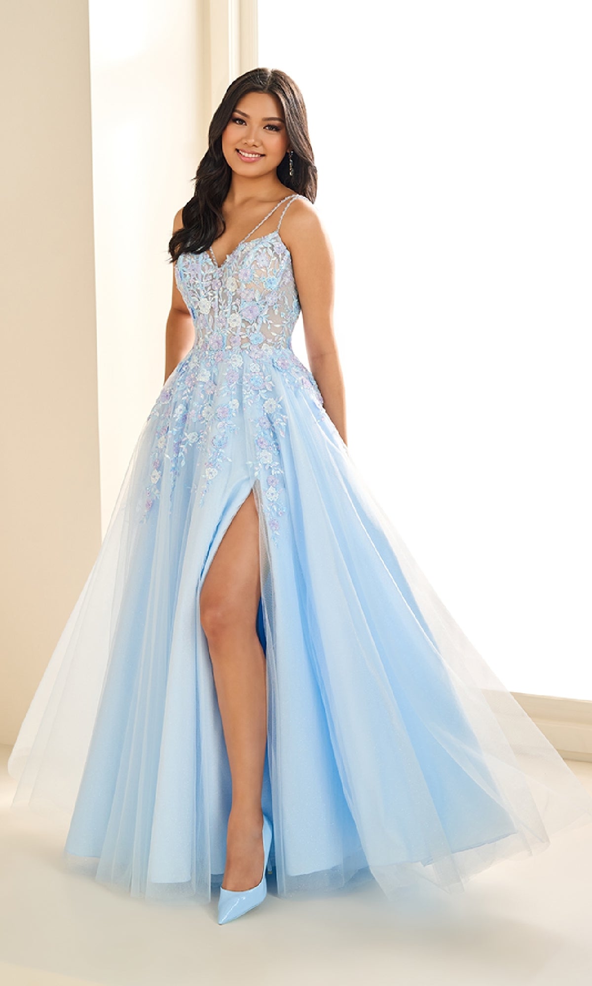 A woman stands indoors wearing the Ellie Wilde EW36079 long prom dress, featuring a light blue ball gown design with a sheer corset bodice, floral embroidery, and a thigh-high slit, paired with matching heels.
